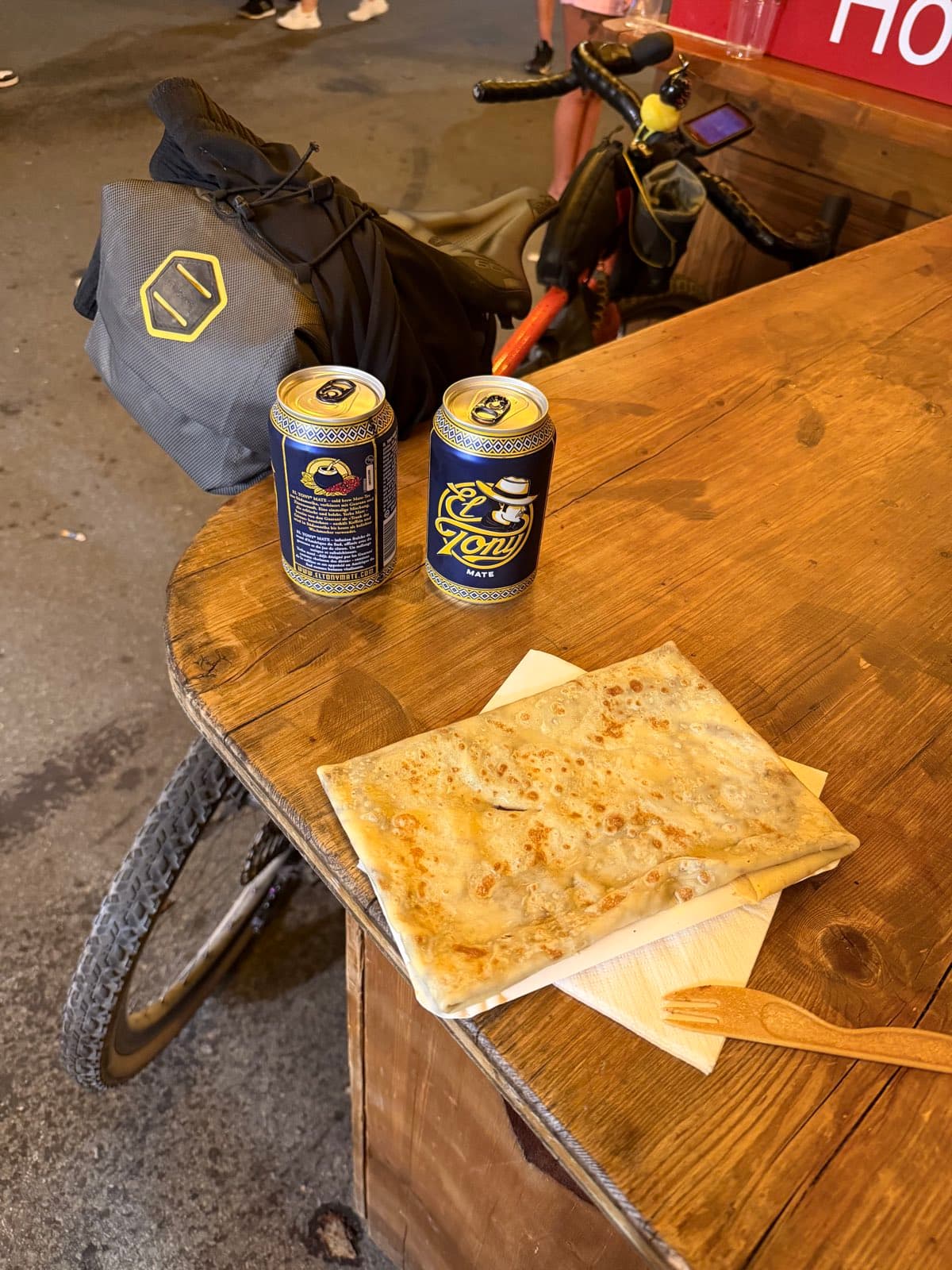 Crêpe auf einem Holztisch neben zwei Dosen Toni Mate, daneben das bepackte Velo