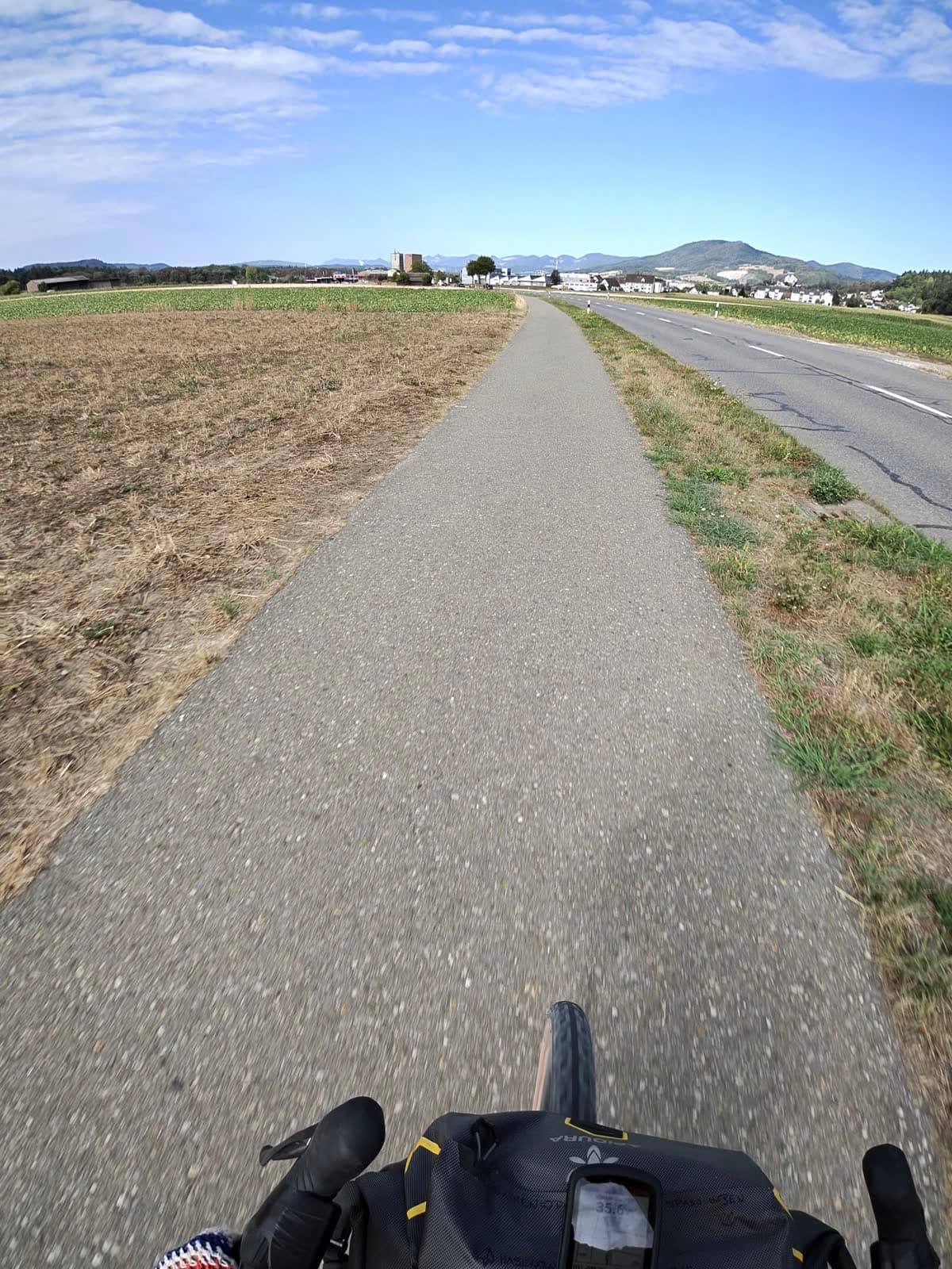 Radweg durch flaches Feld, Jura am Horizont